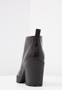 Bota de cuero negro hasta el tobillo con un tacón afilado, superficie texturizada y un tirador; presenta una suela resistente con patrones de tracción.