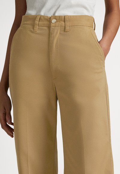 Μπεζ παντελόνι ψηλόμεσο με θηλιές για ζώνη και κούμπωμα με κουμπί, φορεμένο με το ένα χέρι στην τσέπη και λευκό μπλουζάκι μέσα στην παντελόνα.