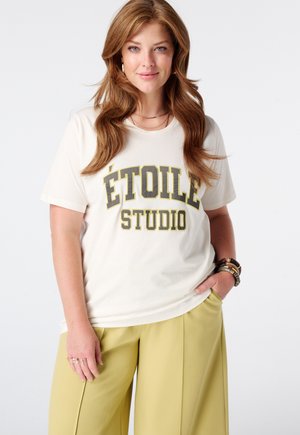 Femme aux longs cheveux bruns portant un t-shirt blanc avec le texte "ÉTOILE STUDIO" et un pantalon jaune à jambes larges, debout avec une main dans la poche.