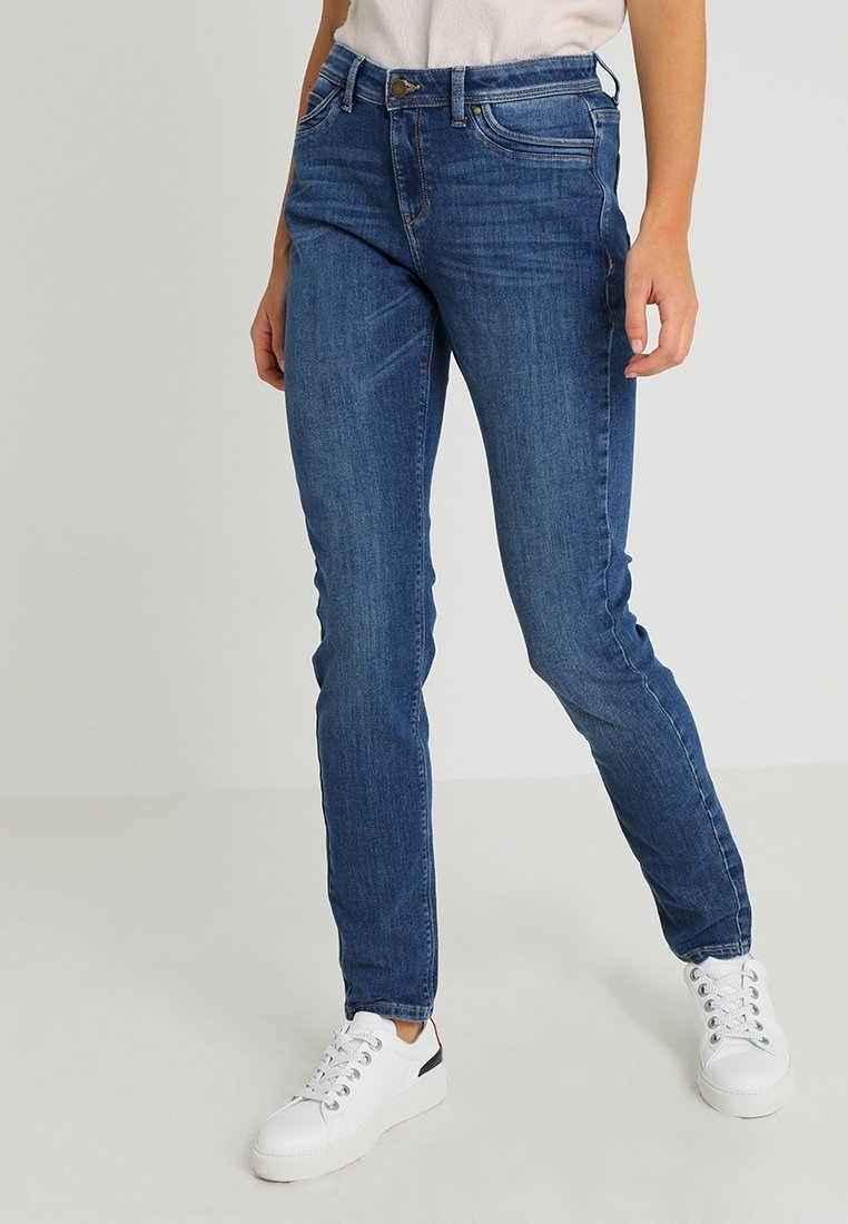 Femme portant un jean slim bleu et des baskets blanches, debout devant un fond clair uni.