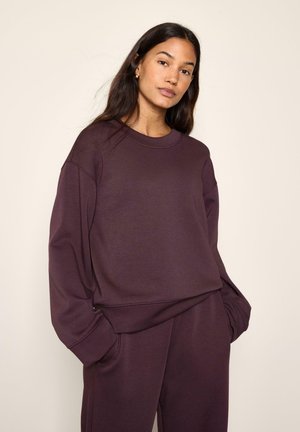 Vrouw met lang donker haar, die een losse donkerpaarse trui en bijpassende broek draagt, staand met handen in de zakken tegen een lichte achtergrond.