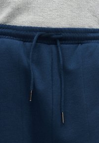 Κοντινό πλάνο σε σκούρο μπλε παντελόνια φόρμας με ελαστική μέση και δεμένη κορδονιέρα με μεταλλικά άκρα, φορεμένα πάνω από ένα ανοιχτό γκρι υφή πουκάμισο.