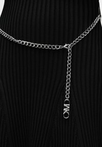 Ασημένια ζώνη αλυσίδα με κρεμαστό κούμπωμα που φέρει τα αρχικά MK, φορεμένη πάνω από μαύρο ριγωτό ύφασμα.