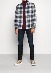Camicia di flanella con un motivo a quadri blu, giallo e bianco; dolcevita bordeaux sotto; jeans scuri; sneakers beige con suole bianche.