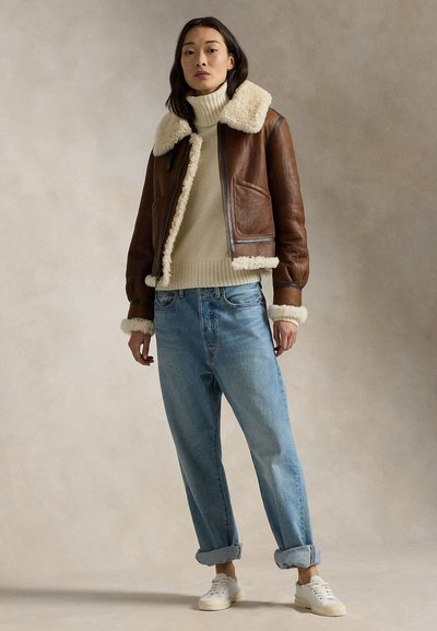 Woman wearing a brown shearling jacket over a cream turtleneck sweater, loose blue jeans rolled at cuffs, and white sneakers, standing.