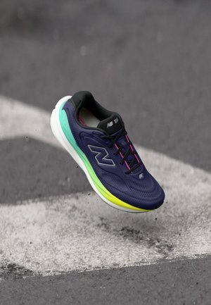 Navy blue and neon green New Balance running shoe suspended above asphalt with white road markings and small pebbles beneath.