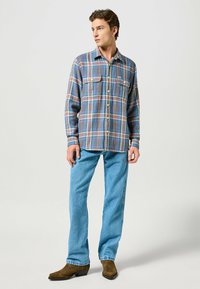 Camisa de cuadros en tonos azules, verdes y naranjas, con dos bolsillos en el pecho, combinada con jeans azul claro y botas marrones.