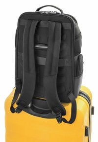 Czarny plecak przymocowany do uchwytu żółtej walizki z twardą powłoką, wyposażonej w wyściełane paski i boczne kieszenie.