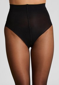 Vrouw in zwarte hoge doorschijnende panty met stevige zwarte slips die heupen en taille bedekken, tegen een grijze achtergrond.