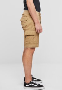 Brune cargoshorts laget av stoff med sidelommer. Har flat front, opprullet kant og diskret søm. Kombinert med svarte joggesko.