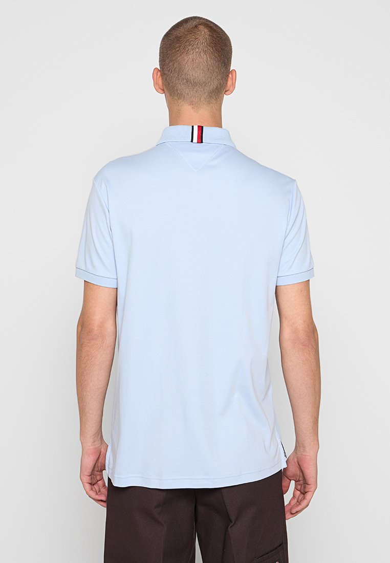 Muž v světle modrém polo tričku s krátkým rukávem, na jehož límečku je červený, bílý a černý pruh, v tmavě hnědých kalhotách, otočený zády.