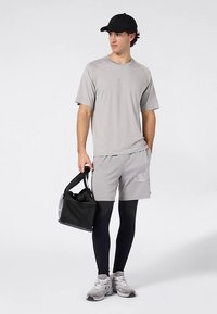 Graues Sport-T-Shirt, graue Shorts mit Logo, schwarze Leggings und schwarze Kappe. Hält eine schwarze Sporttasche. Hellgraue Sportschuhe.