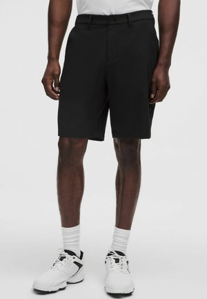 Hombre con pantalones cortos negros hasta la rodilla, calcetines blancos y zapatillas blancas, de pie con los brazos relajados a los costados.