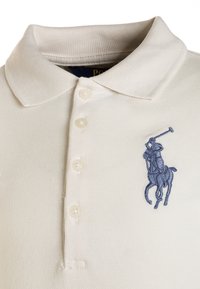 Camisa polo blanca con cuello acanalado y cuatro botones. Un logo de jugador de polo bordado en azul está situado en la parte izquierda del pecho.