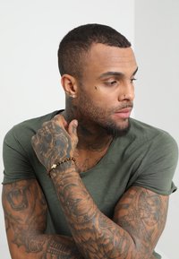 Tricou verde cu mânecă scurtă pe un bărbat cu tatuaje, punând în evidență o brățară din lemn cu un accent în formă de cruce. Textură netedă, croială lejeră.