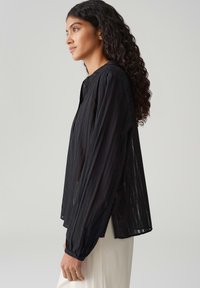 Blusa nera a maniche lunghe con strisce verticali, chiusura con bottoni e polsini arricciati. Tessuto leggero e trasparente con una vestibilità rilassata.