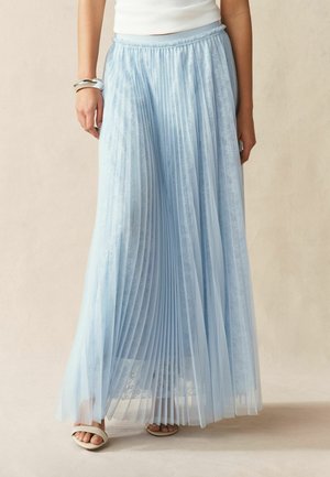 Frau trägt einen hellblauen, plissierten Maxirock mit Spitze, ein weißes ärmelloses Oberteil, weiße Sandalen und ein silbernes Armband am linken Handgelenk.