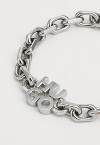 Sidabrinė metalinė grandinėlė su tarpusavyje susijungusiomis saitais ir ryškiu "HUGO" logotipu, apdailinta blizgiu paviršiumi.