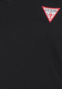 Juodas medvilnės marškinėlis su V formos iškirpte ir raudonu trikampiu logotipo pleistru, ant kurio užrašyta baltais rašmenimis "GUESS" ir klaustukas.