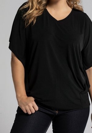 Vrouw met golvend blond haar, gekleed in een loszittende zwarte V-hals top en donkere spijkerbroek, staand met één hand rustend bij haar heup.