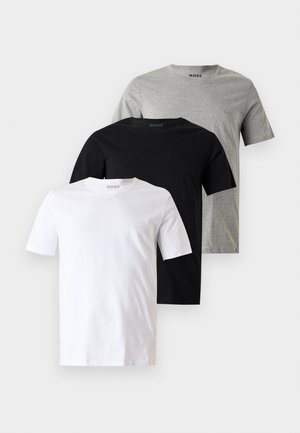 Mężczyzna stoi boso, ubrany w czarny t-shirt z krótkim rękawem i jasno szare spodnie dresowe, z rękami w kieszeniach na jednolitym tle.