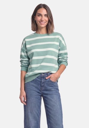 Kvinde med skulderlangt brunt hår iført en grøn- og hvidstribet trøje og blå jeans, smilende med den ene hånd i lommen.