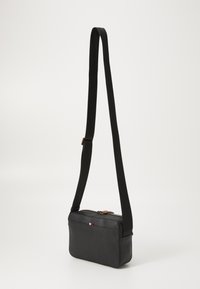 Geantă din piele neagră tip crossbody, cu o formă dreptunghiulară, închidere cu fermoar și un breteau negru. Dispune de un mic logo roșu, alb și albastru.