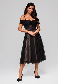 Robe noire décolletée avec un corsage froncé, superposition en tulle à pois, taille cintrée et jupe longueur genoux. Associée à des escarpins noirs ornés de nœuds.