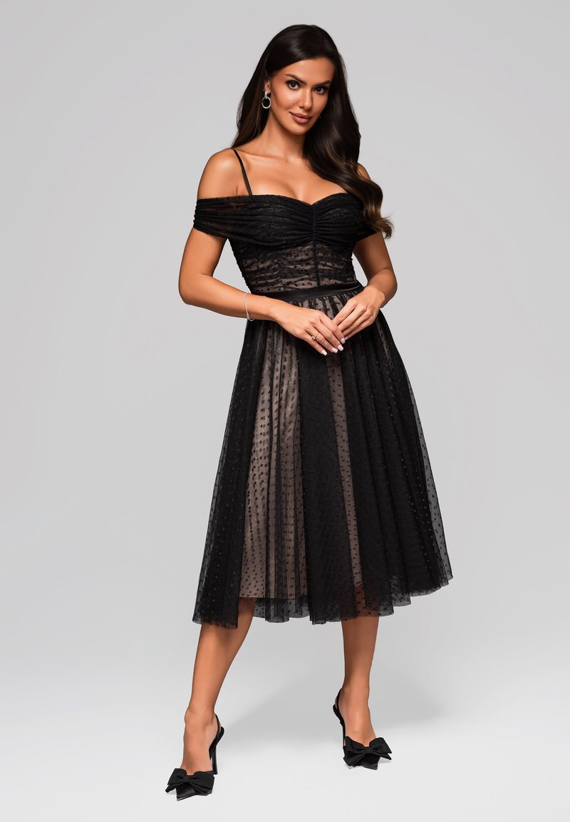 Robe noire décolletée avec un corsage froncé, superposition en tulle à pois, taille cintrée et jupe longueur genoux. Associée à des escarpins noirs ornés de nœuds.