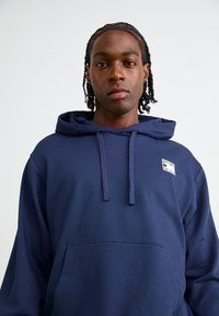 Navy blue hoodie made of soft fabric, featuring a kangaroo pocket, drawstrings, and a small multicolored logo on the chest.