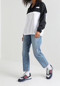 Czarny i biały wiatrówka z ćwierć zamkiem, niebieskie dżinsy, oraz czarne sneakersy New Balance z czerwonymi akcentami i białymi podeszwami.