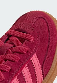 Röda mockaskor med texturerade överlägg, platta snören och gummisula. Designen inkluderar zickzackränder i en ljusare rosa nyans.