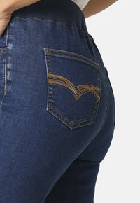 Vaqueros de mezclilla azul con una cinturilla acanalada, un bolsillo trasero con un patrón decorativo de bordado en oro y una silueta ajustada.