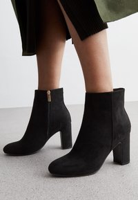 Des bottines en suède noir présentent un talon épais, un bout pointu et une fermeture éclair sur le côté avec un accent doré. La texture semble douce et lisse.