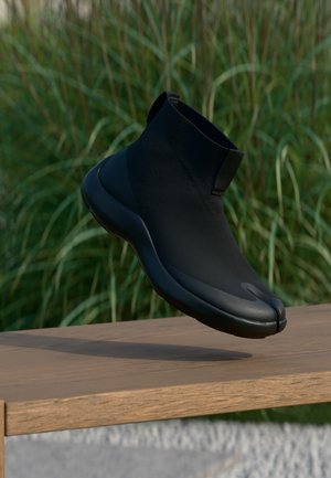 Svarte ankelhøye sokk-stil sko med segmentert tådesign svevende over en trebenk, med grønt gress i bakgrunnen.