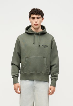 Joven con el pelo corto y rizado que lleva una sudadera con capucha verde oliva con el logo "Service Works" y vaqueros azul claro, de pie frente a un fondo liso.