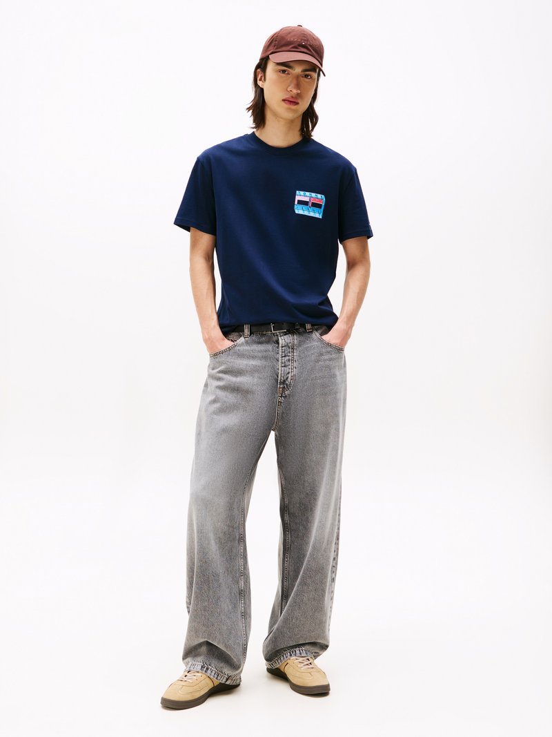 Camiseta navy con un diseño gráfico en el bolsillo, combinada con pantalones vaqueros de pierna ancha en gris desvanecido y zapatillas beiges, complementada con una gorra marrón.