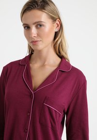 Burgundy pajama top with long sleeves, button closure, contrasting pink piping along the collar and pocket, made from soft fabric.