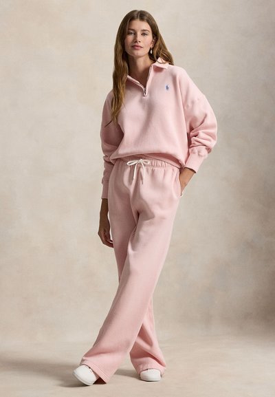 Ανοιχτό ροζ φλις φούτερ με γιακά μισής ζώνης και μικρό μπλε λογότυπο, συνδυασμένο με αντίστοιχο παντελόνι φόρμας με κορδόνι, άνετη γραμμή με φαρδιά πόδια.