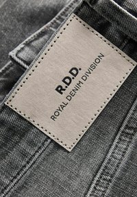 Nahaufnahme von grauen Denim-Jeans mit einem rechteckigen beigen Lederpatch, auf dem "R.D.D. Royal Denim Division" steht.