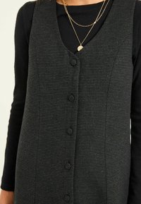 Schwarzes, gestreiftes ärmelloses Kleid mit V-Ausschnitt, das eine Reihe von sechs strukturierten Knöpfen und ein tailliertes Design aufweist. Über einem schwarzen Langarmoberteil getragen.