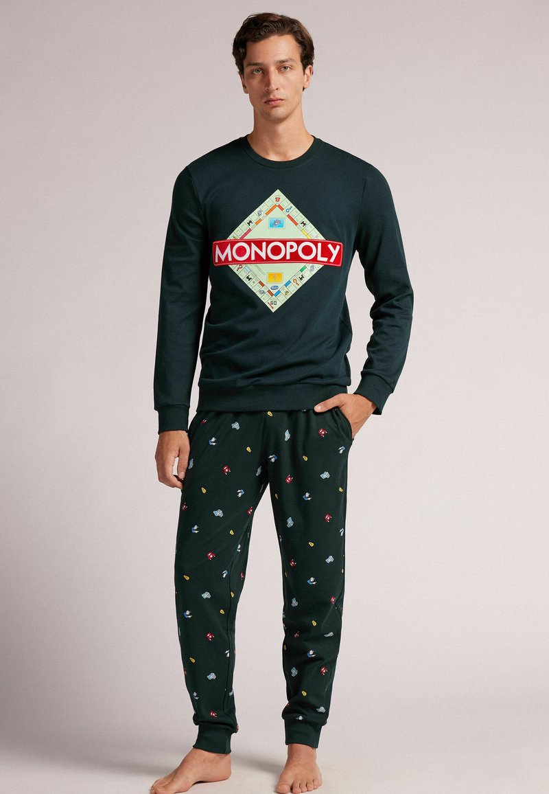Sudadera verde con gráfico de Monopoly en el pecho, combinada con pantalones de pijama verdes que presentan fichas de juego de colores en patrones dispersos.