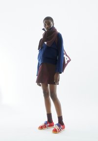 Model trägt eine blaue kurz geschnittene Jacke, einen maronfarbenen Schal, einen maronfarbenen Rock, rote Sneakers mit blauen und weißen Akzenten sowie gestreifte lila Socken.