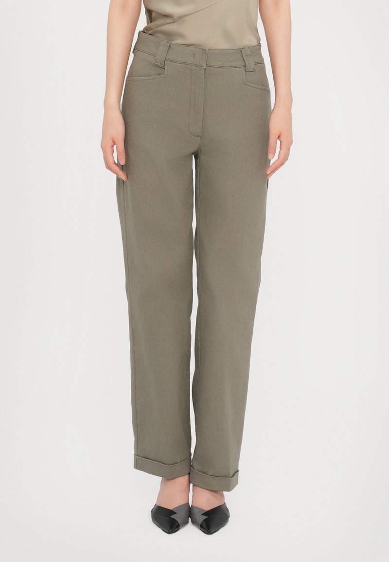 Personne portant un pantalon droit vert olive avec ourlets retroussés et des chaussures noires à bout pointu, debout devant un fond uni.