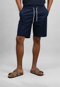 Navy blue shorts with an elastic waistband and drawstring, featuring side pockets. Worn with brown suede slide sandals.