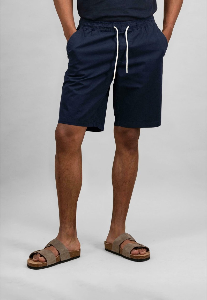 Navy blue shorts with an elastic waistband and drawstring, featuring side pockets. Worn with brown suede slide sandals.