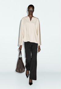 Blouse beige clair ample avec un col en V, pantalons noirs à jambe large et escarpins noirs pointus, portant un sac fourre-tout en cuir marron.