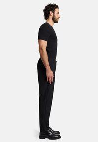 T-shirt nera aderente abbinata a pantaloni neri su misura e scarpe nere lucide. Il modello è in profilo su uno sfondo neutro.