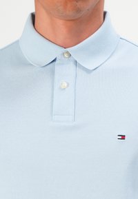 Férfi világoskék galléros pólót visel, amelyen két fehér gomb található, és a mellkasán egy kis téglalap alakú piros, fehér és sötétkék logó látható.