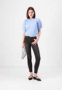 Suéter corto de color azul claro con mangas cortas abullonadas, combinado con jeans ajustados oscuros. La modelo sostiene un bolso de mano plateado con nudo y lleva zapatos planos negros.